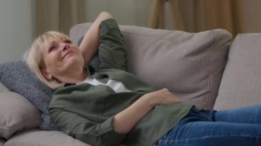 Mutlu, rahat, gülümseyen, yaşlı, beyaz bir kadın kanepeye uzanmış rüya görüyor. Yaşlı bayan, emekli kadın, kanepede dinleniyor.