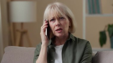 Kafası karışık, yaşlı, cep telefonundan konuşan, kafası karışık yaşlı bir kadın. Evde, kanepe üzerinde olumsuz bir konuşma. Yaşlı, yaşlı bir kadın.