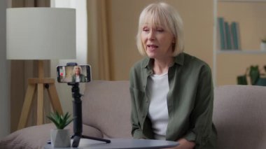 Yaşlı beyaz bir kadın cep telefonuyla video blogu kaydediyor. Vlog sitesinden emekli, akıllı telefon kayıt kanalı olan yaşlı bir kadın. Kanepede oturan telefon tripodu kullanarak nüfuzunu konuşturuyor.