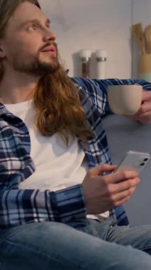 Kafkasyalı rahat bir adam evinde oturmuş çay içiyor elinde cep telefonu erkek ev sahibi sıcak kahvenin tadını çıkarıyor barışçıl adam akıllı telefonu kullanıyor ev kredilerini ev kredisi olarak düşünüyor.