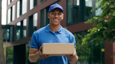 Mutlu gülümseyen Afrikalı Amerikalı kurye elinde paket karton alışveriş siparişi servisi ile dışarıda duruyor. Kamera sevkiyatına bakıyor.
