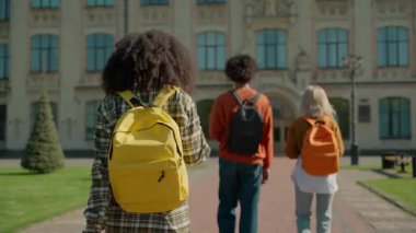 Üç çok ırklı öğrenci arkadaşı Afro-Amerikalı erkek Kafkasyalı kadın kız öğrenci kolej akademisinin önünde yürüyorlar liseye geri dönüp gülümsüyorlar.