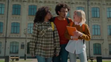 İki çok ırklı kız öğrenci üniversitenin dışında yürüyorlar Afrikalı erkek sarılıyor kızlar konuşuyorlar akademi lisesinde erkek sınıf arkadaşlarını selamlıyor üç mutlu arkadaş