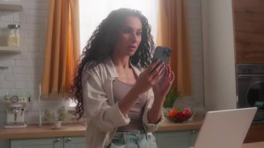 Mutlu İspanyol iş kadını zaferi kutluyor cep telefonu ev mutfağı Latin kadın iş kadını internette başarılı bir başarı mesajı bağırıyor bilgisayarlı akıllı telefon başarılı kız