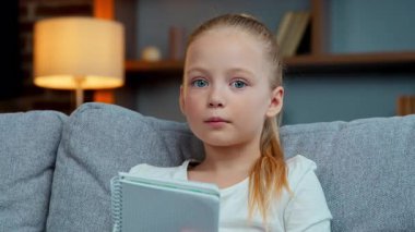 Kafkasyalı küçük kız çocuğu kitap okuyor ev videolarına bakıyor e-öğrenim metni okuyor kanepe üzerinde ders kitabı tutuyor çevrimiçi konferans eğitimi öğreniyor.