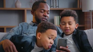 Cep telefonuyla gülümseyen Afro-Amerikan mutlu aile fotoğraf çeken baba suratını buruşturan çocuklar akıllı telefondan bakan çocuklar evde video kaydı yapmaya çalışan komik maske takan çocuklar.