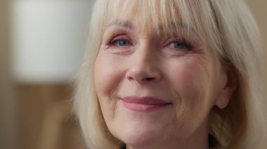 Düşünceli yaşlı beyaz kadın kameraya bakıp gülümsüyor mutlu gülüyor mutlu gülümsüyor dişlek, dişlek, beyaz dişli yaşlı bayan büyükanne içeride olumlu duygularla gülüyor.