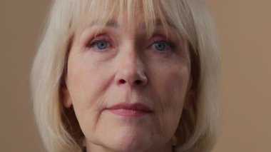 Kapalı portre, yaşlı beyaz buruşuk kadın, sakin ol. Kameraya ciddi ciddi bak. Yetişkin, gri saçlı, yaşlı, yaşlı, yaşlı kadın, emekli büyükanne, vesikalık büyükanne bej renkli stüdyoya bakıyor.