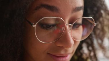 Aşırı yakın plan Afro-Amerikan kadın gülümsüyor, evde çalışıyor, etnik kökenli bayan öğrenci öğreniyor, gözlük takıyor, üniversite akademisinde öğrenim görüyor ve gülümsüyor.