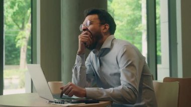 Hintli etnik iş adamı, sabahın erken saatlerinde dizüstü bilgisayar yazarken Arap kahvesini kafeteryada esnetirken, esnetirken, erkek adam kahve içerken bilgisayar stresi, uykusuzluk ve yorgunluk problemleri kullanarak masadaki ziyaretçiyi rahatsız ediyor.