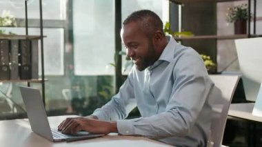 Kaygısız Afrikalı Amerikalı iş adamı ofiste dizüstü bilgisayarda dans ederken müzik dinliyor olgun sesli iş adamı eğleniyor 40 'lı yaşlardaki yönetici bilgisayar kullanarak iyi iş çıkarmayı kutluyor.