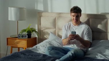 Gülümseyen beyaz adam yatak odasında uzanmış akıllı telefon kullanıyor kaygısız erkek sohbet ediyor gülüyor sosyal medya cep telefonu ağına gülüyor evde rahatça rahatlıyor.