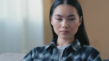 Düşünceli ciddi Asyalı kadın, evinde oturmuş kameraya bakıyor. Sakin, odaklanmış, ifadeye odaklanmış, Koreli, Çinli kadın, ev hanımı, düşünceli, genç, kendine güvenen, barışçıl bakışlar atıyor.