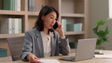 Meşgul Asyalı iş kadını cep telefonu incelemesi belgelerini bilgisayarda ararken Çinli iş kadını Koreli bayan Japon işvereni ofiste akıllı telefonlardan konuşuyor.