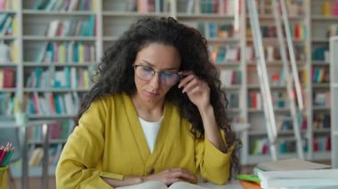 Depresyonda olan Latin bir kadın elinde baş ağrısı ağrısı ile kütüphanede ders kitabı okuyor kız öğrenci öğrenim sınavı okuyor aşırı çalışan migren üniversitesi öğrencisi kitapçı kız okuyor.