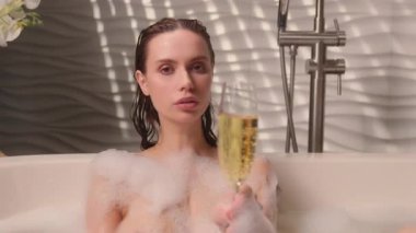 Köpüklü köpüklü bir köpük banyosunda oturan genç beyaz kadın elinde bir kadeh şampanya, zarif bir bayan kız. Sıcak suya doğru içecek koyuyor. Lüks, sakin, rafine kaplıca atmosferi.