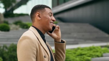 Şehirde cep telefonuyla konuşan mutlu Afro-Amerikalı iş adamı olumlu gülümseyen girişimci yatırımcı erkek akıllı telefon görüşmeleri kentsel pazarlamacı iş adamını arıyor.