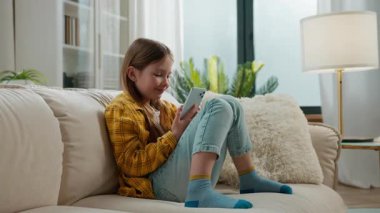 Gadget bağımlısı küçük beyaz kız cep telefonunu evde kullanıyor mutlu gülümseyen akıllı telefon teknolojisi kanepe kızının evinde sosyal medyada komik videolar seyrediyor gülümsüyor.