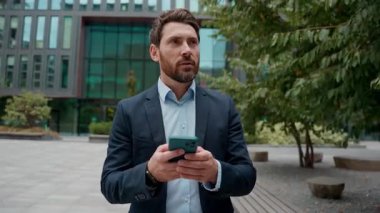 Kafkasyalı mutlu yetişkin iş adamı şehirde cep telefonu uygulaması kullanarak yürüyor orta yaşlı sohbet eden akıllı telefon profesyonel iş adamı iletişimi erkek teknolojisi açık hava