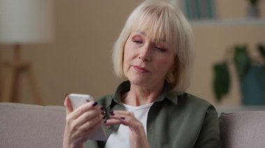 Mutlu gülümseyen Kafkasyalı yaşlı kadın cep telefonu gülüşü kaygısız kaydırmalı akıllı telefon uygulaması kullanıyor koltukta oturan olgun yaşlı bayan cep telefonu dokunmatik internet alışverişi e-ticareti yapıyor.