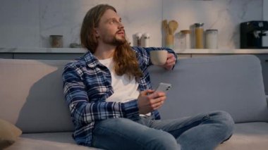 Kafkasyalı rahat bir adam evinde oturmuş çay içiyor elinde cep telefonu erkek ev sahibi sıcak kahvenin tadını çıkarıyor barışçıl adam akıllı telefonu kullanıyor ev kredilerini ev kredisi olarak düşünüyor.