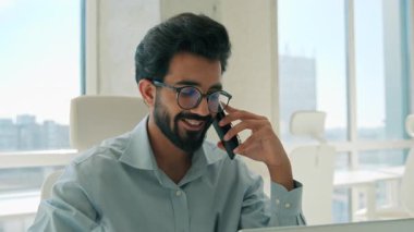Sırıtkan Arap Hintli iş adamı cep telefonuyla konuşurken laptop ofisi erkek iş adamı etnik erkek girişimci işveren müşteriye danışıyor cep telefonu görüşmesi yapıyor.