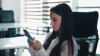 Ofiste cep telefonu kullanarak rahatlayan Kafkasyalı ciddi iş kadını. Mola veren iş kadını. Akıllı telefona göz gezdiriyor.