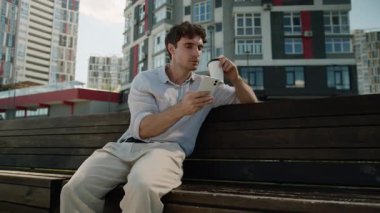 Memnuniyetle gülümseyen Kafkasyalı iş adamı cep telefonu sosyal medya aleti bağımlısı iş adamı bankta oturan adam şehirdeki internet sitesinde gezinen internet sitesini içiyor.