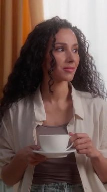 Düşünceli İspanyol kadın elinde fincanla kameraya dönüp gülümsüyor. Latin beyaz kadın ev hanımı. Sıcak aromalı kahve içiyor.