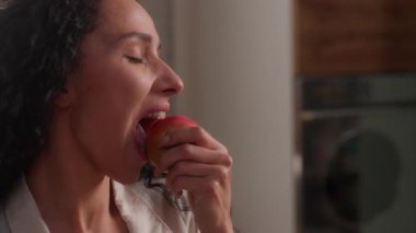 Mutlu, gülümseyen Latin bir kadın, mutfak mutfağında elma göz kırpması yiyor. Beyaz, kadın, vejetaryen, meyve yemesi sağlıklı beslenme diyeti vitamini, ev ortamında sağlıklı yiyecek yiyor.
