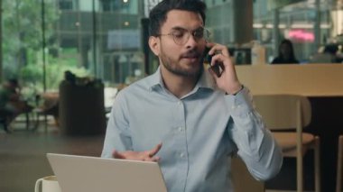 Sinirli agresif, hayal kırıklığına uğramış Hintli işadamı kafede laptopla cep telefonuyla bağırarak konuşuyor öfkeli, sinirli iş adamı, akıllı telefon sorununu tartışıyor.