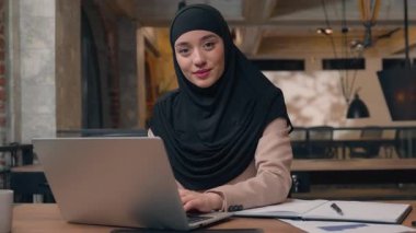 tesettürlü mutlu iş kadını ofis işkadınını davet ediyor jest işe alım daveti kameraya gülümseyen Arap kız müslüman bayan çalışan dizüstü bilgisayara bakıyor buraya gelin