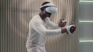 Afro-Amerikalı kaygısız erkek, 3 boyutlu sanal gerçeklik kaskı takan VR gözlük takıyor, simülasyon oynuyorlar modern metaevren oyunu oynuyorlar gelecekteki beyaz stüdyoda silah kontrolü yapıyormuş gibi davranıyorlar.