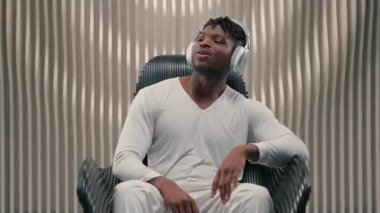 Futurizm Afrikalı Amerikalı erkek adam beyaz fütüristik odada kapalı mekanda siber uzay modern teknoloji kablosuz kulaklıklarda müzik dinlerken sandalye dansında dinlenen şarkı sesleri dinlemenin tadını çıkarın.