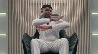 Afro-Amerikalı adam kulaklıkla müzik dinliyor, dans ediyor, koltukta dinleniyor, dans ediyor, el sallıyor, dans ediyor, kulaklık takıyor, fütürizm uygulamasında şarkı dinliyor.