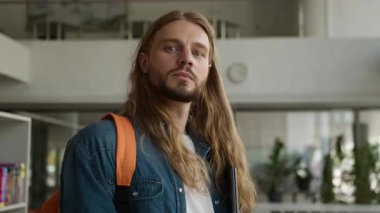 Kafkasyalı öğrenci erkek, üniversite akademisi koridorunda tasasız, tasasız, elinde dizüstü bilgisayarla kameraya bakıp mutlu bir şekilde gülümseyen ve öğrenim başarısı eğitimi alan biri.