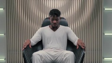 Kendine güvenen, Afro-Amerikalı, fütüristik uzayda sandalyede oturan beyaz odada gülümseyen siber erkeğe bakıp fütürizm stüdyosunda rahatlayan neşeli, tasasız bir şekilde oturuyor.