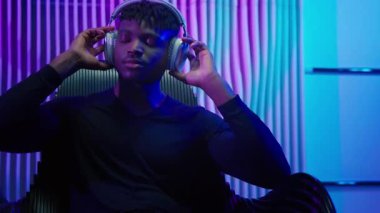 Fütüristik odada Afro-Amerikalı bir adam kulaklıkla müzik dinliyor DJ dans ediyor koyu mavi neon fütürizm alanında kafa dansı yapıyor sandalyeye oturmuş dinleniyor erkek adam molası verip şarkı dinliyor.