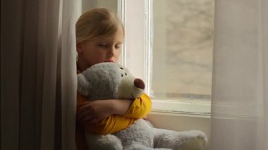 Stresli küçük kız çocuğu pencereye bakıyor oyuncak ayıya sarılıyor evde yalnızlık çekiyor evde yalnızlık çekiyor kederli bir çocukluk çocuğu evde yalnız başına zihinsel sağlık psikolojisini benimsiyor.