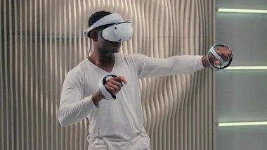 Afro-Amerikan fütürist adam sanal gerçekliği kullanarak VR oyunundan zevk alıyor. Fütürizm stüdyosu oyuncusu erkek oyuncu, eğlence amaçlı sanal ortam için kask tutucu kontrolörleri ile çevrimiçi oynuyor.