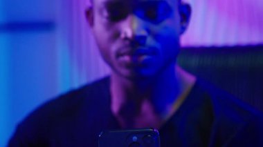 Afrikalı Amerikalı adam cep telefonuyla kazanmak için bağırmaktan mutlu. Karanlık neon fütüristik uzay tasasız kazananı erkek akıllı telefonla zaferi kutluyor. Fütürizm teknolojisi elde ediyor.