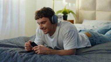Neşeli Kafkasyalı adam evde yatarken kulaklık takıyor. Akıllı telefon kullanıyor. Dans ediyor, dans ediyor, mutlu erkek.