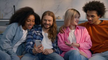 Çok ırklı arkadaşlar erkekler kadınlar evdeki koltukta yemek siparişi veriyorlar Afrikalı beyaz kızlar erkekler akıllı telefon sosyal medya çok ırklı arkadaşlığı kapalı mekanda tartışıyorlar.