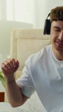 Etkin, enerjik, mutlu, beyaz, müzik kulaklığı dinleyen, yatak odasında dans eden, sabah çalma listesinin keyfini çıkaran, cep telefonlu, rahat, evde dinlenen ve eğlenen genç bir adam.