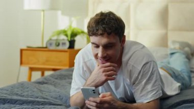 Beyaz adam yatakta uzanmış gülümsüyor. Yatak odasında akıllı telefon kullanıyor. Rahatlamış erkek mesajları atıyor. İnternette sohbet ediyor, sosyal medya internet sahibi evde cep telefonu keyfi yapıyor.