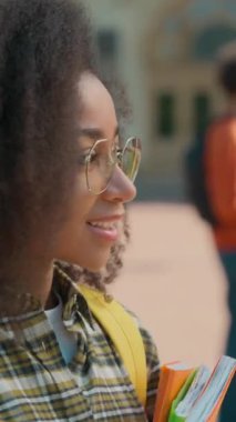 Kameraya bakıp gülümseyen Afro-Amerikan öğrenci elinde kitaplarla üniversite arka plan öğrencileri gözlüklü pozitif etnik kadın portresi konuşuyor.