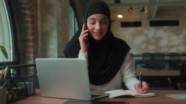 Neşeli gülümseyen cep telefonlu Müslüman kadın tesettürlü iş kadını Arap kadın çoklu iş kadını yönetici ofis ajansında çalışan dizüstü bilgisayar görünümlü iş kadını proje düzenini tartışıyor