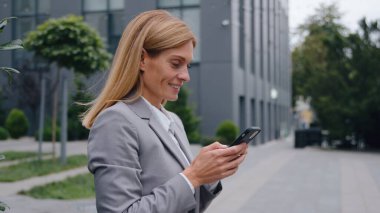 Kafkasyalı mutlu orta yaşlı iş kadını telefonuyla sohbet ediyor şehir merkezinde cep telefonlu bir iş kadını ve cep telefonuyla e-posta yazarken gülümsüyor.