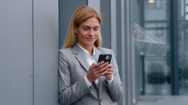 Gülümseyen Kafkasyalı orta yaşlı iş kadını cep telefonu tutuyor şirket binasının duvarına yaslanıyor gülümsüyor mutlu 40 'ların iş kadını şehirde e-posta sohbet akıllı telefonu kontrol ediyor.