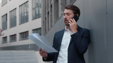 Kafkasyalı orta yaşlı iş adamı şehirde telefon tutan kağıtlardan bahsediyor ofis duvarlarına yaslanıyor yetişkin bir erkek işadamı şehir merkezinde cep telefonuyla konuşuyor ve belgeleri tartışıyor.